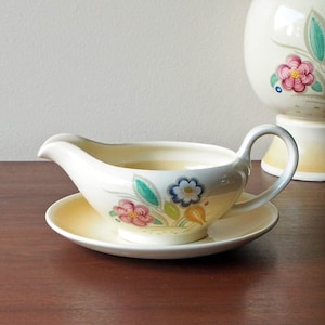 May include: A white ceramic gravy boat with a floral design in shades of pink, blue, green, and yellow. The boat is on a matching saucer.