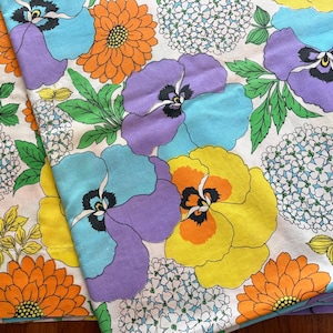 May include: A fabric featuring a vibrant floral pattern on a white background. The design includes large purple and yellow pansies, bright orange chrysanthemums, and clusters of small white hydrangea blossoms with green leaves throughout the print.