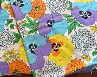 May include: A fabric featuring a vibrant floral pattern on a white background. The design includes large purple and yellow pansies, bright orange chrysanthemums, and clusters of small white hydrangea blossoms with green leaves throughout the print.