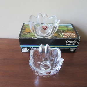 May include: Two clear glass candle holders with a floral design. The candle holders are shaped like a flower with curved petals. The box behind the candle holders has the text "Orrefors Sweden".