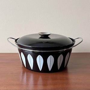 Könnte beinhalten: Ein schwarzer Emaille-Kochtopf mit Deckel und silbernen Griffen. Die Schüssel hat ein weißes Blättermuster um den Boden. Der Deckel hat einen silbernen Knopf. Ein Vintage-Küchenartikel.
