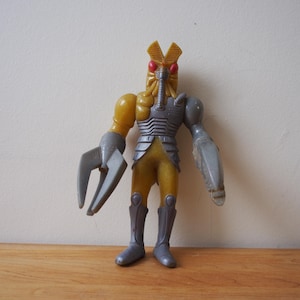 May include: A yellow and silver action figure of a humanoid insect with red eyes and a long snout. The figure has two large claws on each hand.