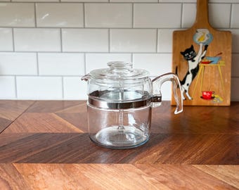 Peut inclure: Une théière en verre transparent dotée d'un corps cylindrique, d'un couvercle en verre assorti et d'une poignée en verre robuste. Une bande métallique argentée entoure la partie supérieure du récipient, fixant la poignée et contrastant avec le verre.