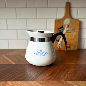 Könnte beinhalten: Weiße Vintage-Kaffeekaraffe mit schwarzem Griff und silbernem Band. Die Karaffe hat ein blaues Blumendesign. Im Hintergrund befindet sich ein Holzschneidebrett mit einer Katzenillustration.