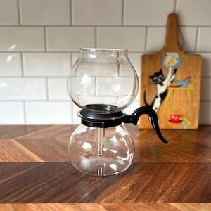 Könnte beinhalten: Ein klarer Glas-Siphon-Kaffeebereiter mit schwarzem Griff und Sockel. Der Kaffeebereiter steht auf einer Holzoberfläche. Im Hintergrund befindet sich ein dekoratives Holzschneidebrett mit einer Katzenillustration.