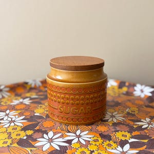 May include: A cylindrical ceramic sugar canister with a warm orange and brown gradient glaze. The exterior features a repeating embossed floral and circular pattern with the word SUGAR centred on the front. It has a flat, circular wooden lid.