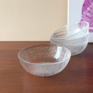 以下が含まれることがあります： テクスチャ加工された垂直パターンが施された透明なガラスボウル。1つのボウルに焦点が当てられ、その背後に3つのボウルのスタックがあります。ボウルは濃い木製の表面にあり、背景には額入りのアートワークがあります。