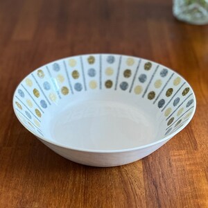 Könnte beinhalten: Eine weiße Keramikschüssel mit einem geometrischen Muster aus goldenen, grauen und weißen Punkten und Linien. Die Schüssel steht auf einer Holzoberfläche.