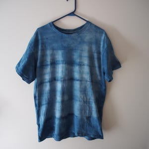 May include: A blue tie-dye t-shirt hanging on a blue hanger against a white wall. The short-sleeved shirt has a round neckline and a horizontal striped pattern in varying shades of blue.