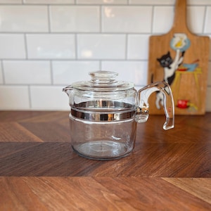 May include: A clear glass coffee pot with a silver band and handle. The pot has a lid and a spout for pouring. It sits on a wooden surface with a herringbone pattern. A decorative wooden cutting board with a cat illustration is in the background.
