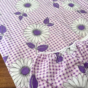 May include: A fitted sheet featuring a white daisy floral design set against a purple and white gingham backdrop. The daisies have white petals and purple centres. The sheet has a white, ruffled edge.