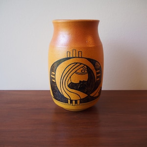 May include: A ceramic vase with a yellow and orange gradient. The vase features a black graphic of a woman's profile within a circular design. The vase is cylindrical with a slightly flared top, and it sits on a wooden surface.