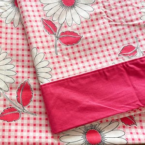 Peut inclure: Un textile en tissu présentant un motif à carreaux vichy rose et blanc, orné de grandes fleurs de marguerites blanches avec des cœurs roses et des feuilles roses. Une bordure en tissu rose uni est repliée sur le haut du matériau à motifs.