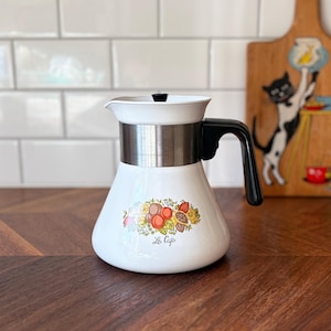 May include: A white enamel coffee pot with a black handle and lid knob. The pot features a stainless steel band around the upper section and a colourful fruit and nut graphic on the front with the script text Le Cafe printed below the design.