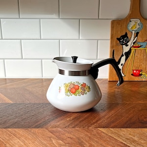 Könnte beinhalten: Weiße Vintage-Teekanne mit schwarzem Griff und Deckel, mit einem farbenfrohen Obst- und Blumendesign. Die Teekanne steht auf einer Holzoberfläche, mit einem dekorativen Holzschneidebrett im Hintergrund.