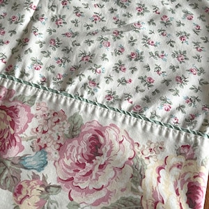 May include: Floral fabric with a pattern of large pink roses and smaller pink and blue flowers on a white backdrop. A green trim divides the two patterns. Suitable for bedding or home decor.