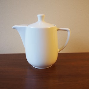 May include: A white ceramic teapot with a lid and handle. The teapot has a curved spout and a rounded body. The handle is a simple, rectangular shape. The teapot is sitting on a dark brown surface.