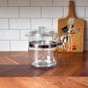 May include: A clear glass coffee percolator with a silver band and a glass lid. The percolator sits on a wooden surface with a decorative cutting board in the background. The cutting board has a painted cat design.