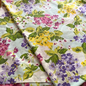 May include: Pale blue fabric with a floral design of yellow, purple, and pink flowers and green leaves. The fabric is folded, revealing the pattern's repeat. The flowers are grouped, creating a bright and colourful design.
