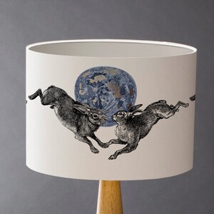 May include: Cream-colored lampshade featuring a detailed illustration of two hares leaping towards each other, set against a blue and gold celestial sphere. The lampshade is mounted on a wooden base, creating a warm, inviting glow.