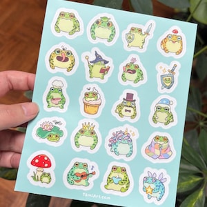 Könnte beinhalten: Ein Bogen mit Aufklebern mit Cartoon-Fröschen in verschiedenen Outfits. Die Frösche sind grün, gelb und orange, einige tragen Hüte, halten Gegenstände oder spielen Instrumente. Der Hintergrund ist hellblau. Die Website TamiArt.com ist sichtbar.
