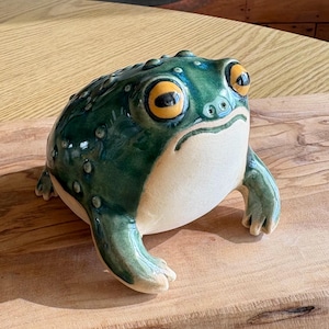 May include: A ceramic frog figurine with a glossy green finish and a cream-colored belly. The frog has large, yellow-ringed eyes and is posed on a wooden surface. The figurine is a decorative item.