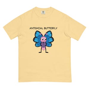 May include: A pale yellow short-sleeve t-shirt featuring a graphic of a pink butterfly character wearing a purple hoodie and blue headphones. The butterfly has blue wings with black and white details. The text ANTISOCIAL BUTTERFLY is printed above.