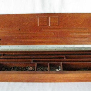 May include: Open wooden tool chest with a hinged lid. The chest is divided into compartments of various sizes, filled with small metal hardware. The wood has a warm, reddish-brown tone, and the lid is slightly ajar.