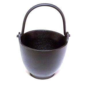 May include: A small, black cast iron pot with a handle. The pot is empty and has a smooth, rounded bottom.
