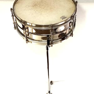 May include: A vintage chrome snare drum with a white drumhead, mounted on a chrome stand. The drum has a shiny, reflective surface and is supported by a tripod base. The drumhead has a Remo logo.