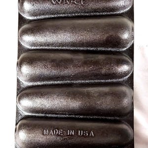 May include: A vintage cast iron hot dog mold with eight individual slots. The mold is black and has the words "Wagner Ware" and "Made in USA" embossed on it.