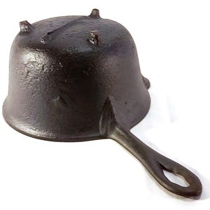 May include: A small, cast iron skillet with a single handle and a rounded, bell-shaped cooking surface. The skillet is dark brown and has three small feet on the top. The handle has a hole for hanging.