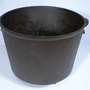 May include: A dark brown cast iron pot with a wide opening and two side handles. The pot has a textured surface and sits on three small feet. This cooking pot is suitable for outdoor use.