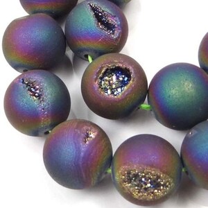 May include: A strand of iridescent agate beads with a druzy finish. The beads are round and have a dark purple, blue, and green color with a metallic sheen. Each bead has a natural crystal cavity filled with sparkling crystals.