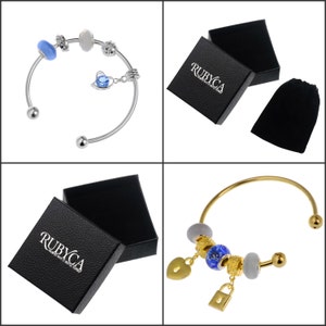 Op de afbeelding: Twee charmbレスレットen in zilver en goud met blauwe en witte kralen, hart- en slotbedels, en een zwarte fluwelen zak en doos met het logo "RUBYCA" erop gedrukt.