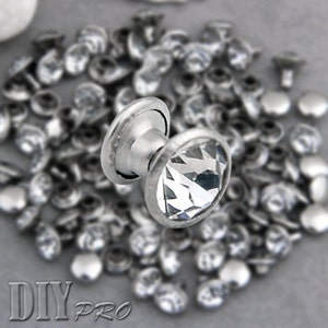 May include: A close-up of a silver metal rivet with a clear crystal top. The rivet is surrounded by a pile of other silver rivets.