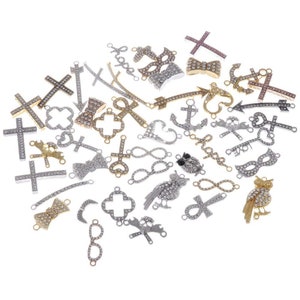 Op de afbeelding: Assortiment van zilveren en goudkleurige bedels met strassaccenten. Bedels omvatten kruisen, ankers, oneindigheidssymbolen, uilen en pijlen.