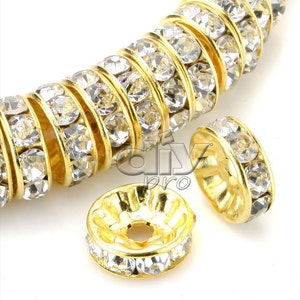 Puede incluir: Cuentas espaciadoras de metal dorado con brillantes pedrería transparente. Las cuentas son redondas con un agujero central y están dispuestas en una fila. También se ven dos cuentas sueltas. La imagen está bien iluminada y muestra los detalles.