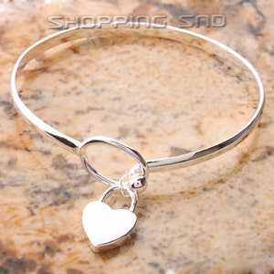 May include: A silver bangle bracelet with a heart-shaped charm. The charm is attached to the bracelet with a small ring and a ball.