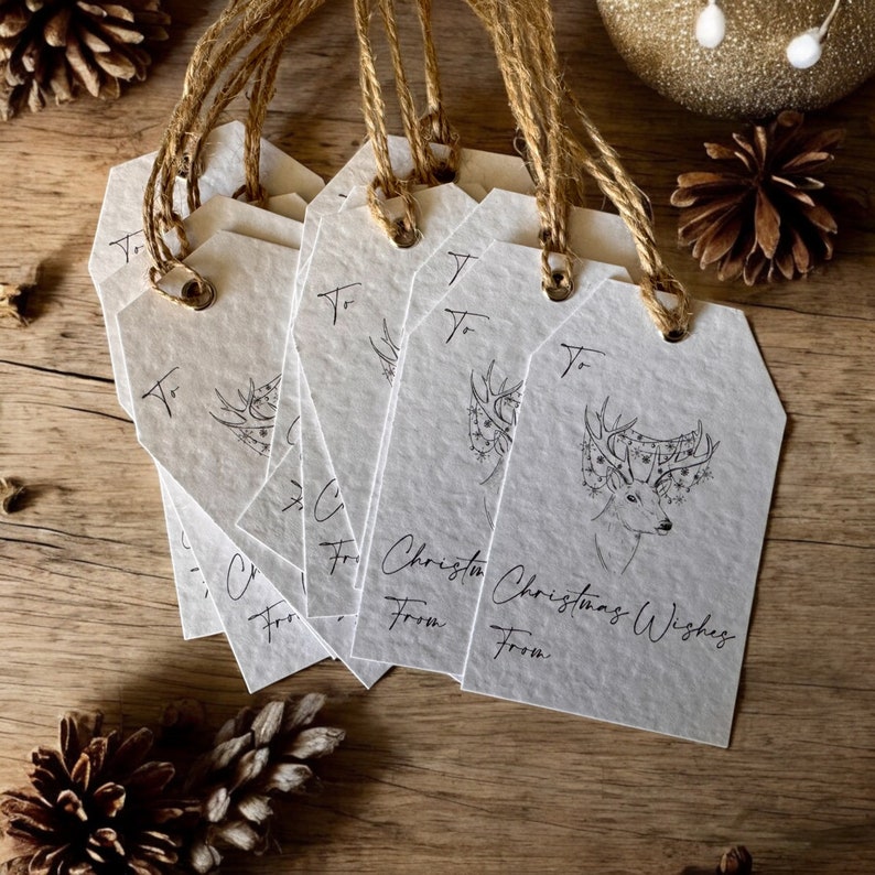 May include: A stack of white gift tags with a black and white illustration of a deer with a Christmas tree on its antlers. The tags have the text "To" and "Christmas Wishes" written on them.
