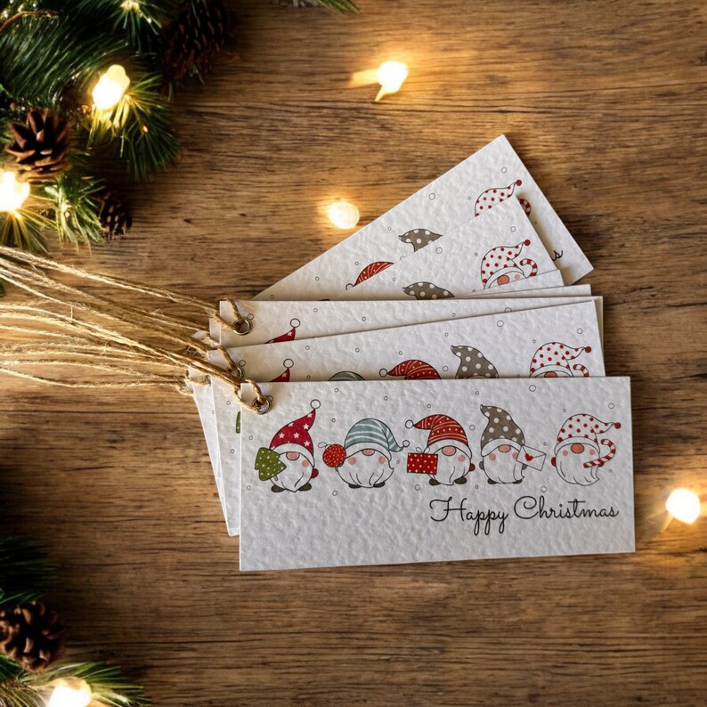 May include: Set of six Christmas gift tags featuring a line of gnomes with red, green, and white hats. The tags are white with a speckled texture and have a brown twine string. The text "Happy Christmas" is printed on the bottom of the tags.