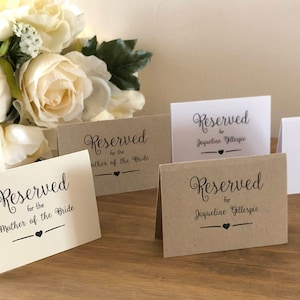 May include: Five place cards for a wedding reception. Two are white with black text that reads "Reserved for the Mother of the Bride". Two are brown with black text that reads "Reserved for Jacqueline Gillespie". One is white with black text that reads "Reserved for Jacqueline Gillespie".