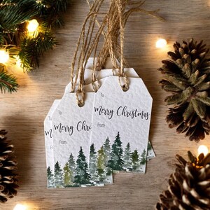 May include: Set of six white gift tags with a watercolor illustration of a forest scene. The tags have the text "Merry Christmas" and "To" and "From" printed on them. The tags are tied with twine.