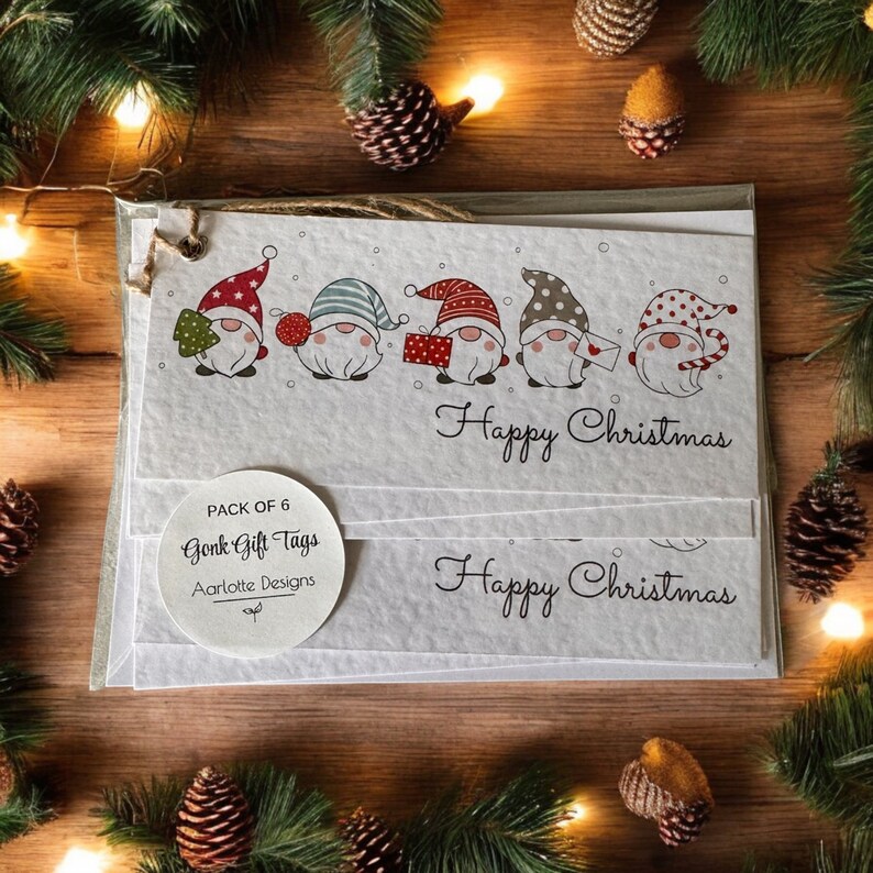 May include: A set of five white gift tags with a gnome design. Each tag features a different gnome with a red hat, a green hat, a blue hat, a brown hat, and a red and white polka dot hat. The tags are decorated with snowflakes and the text "Happy Christmas". The tags are packaged in a white circle with the text "Pack of 5 Gnome Gift Tags Aarlote Designs".