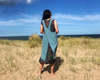 May include: A woman wearing a light blue denim apron dress with a criss-cross back detail. She is standing on a sandy beach with tall grass in the foreground and a blue ocean in the background.