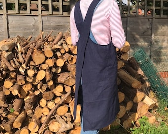 May include: A navy blue linen apron with criss-cross straps in the back. The apron is worn over a pink and white striped shirt and blue jeans.