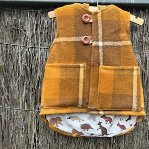 May include: A yellow and brown plaid waistcoat with a hood and two wooden buttons. The waistcoat has two pockets and a lining with a pattern of small animals.