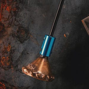 Puede incluir: Una herramienta de precisión con un eje negro y un mango y base azules. El mango tiene un diseño texturizado. La cabeza de la herramienta es de color latón con un borde decorativo. La herramienta se encuentra sobre un fondo oscuro y texturizado.