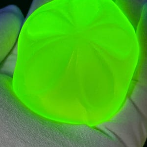 May include: A glowing, neon green, rounded object with a textured surface. The object has a unique, organic shape with subtle patterns. The bright color and smooth curves create a striking visual effect. The object is held in a white fabric.