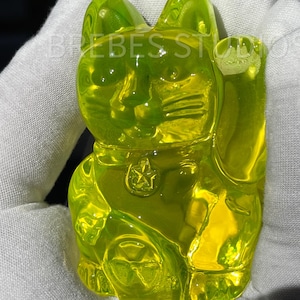Puede incluir: Una figura de gato de la suerte en color amarillo neón translúcido con la pata izquierda levantada, un collar con un colgante en forma de estrella y un emblema de moneda circular en el pecho. El gato tiene rasgos faciales moldeados.